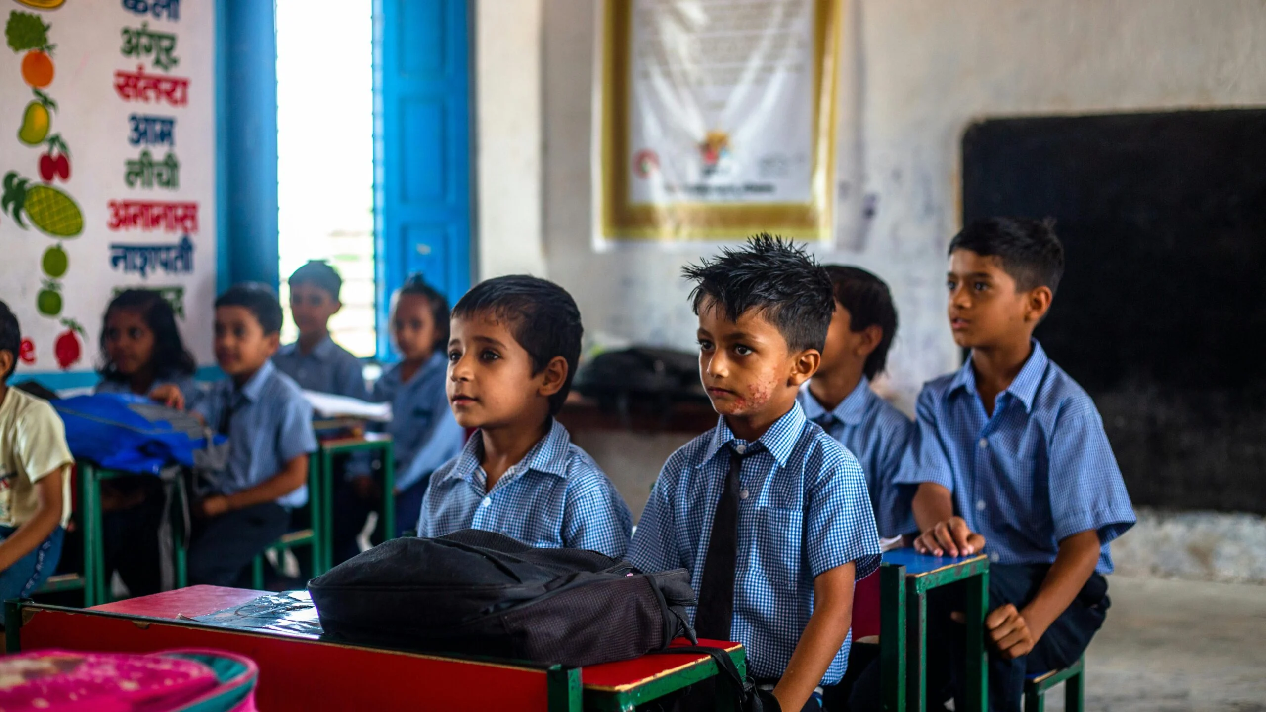 Schoolchildren in blue uniforms sitting at desks in a classroom with a Hindi alphabet chart on the wall