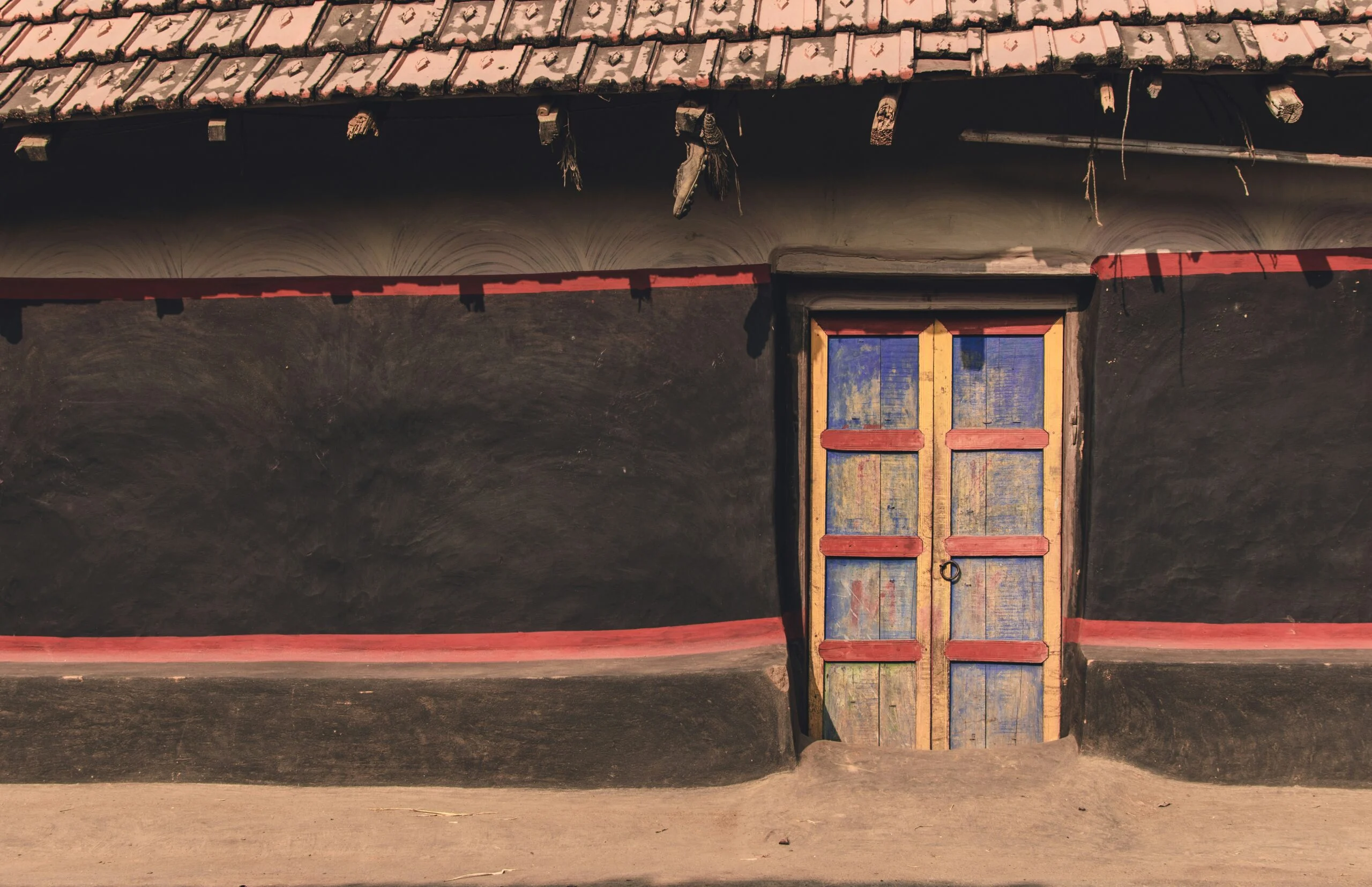 Closed blue and yellow wooden door in a dark mud-plastered wall under a tiled roof in a village