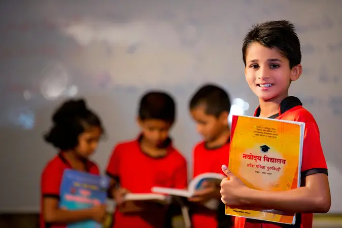 Smiling schoolboy in a red uniform holding an exam guide book, with classmates reading behind him