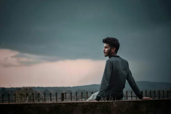 Young man sitting on a low wall at dusk, looking away under a dark cloudy sky