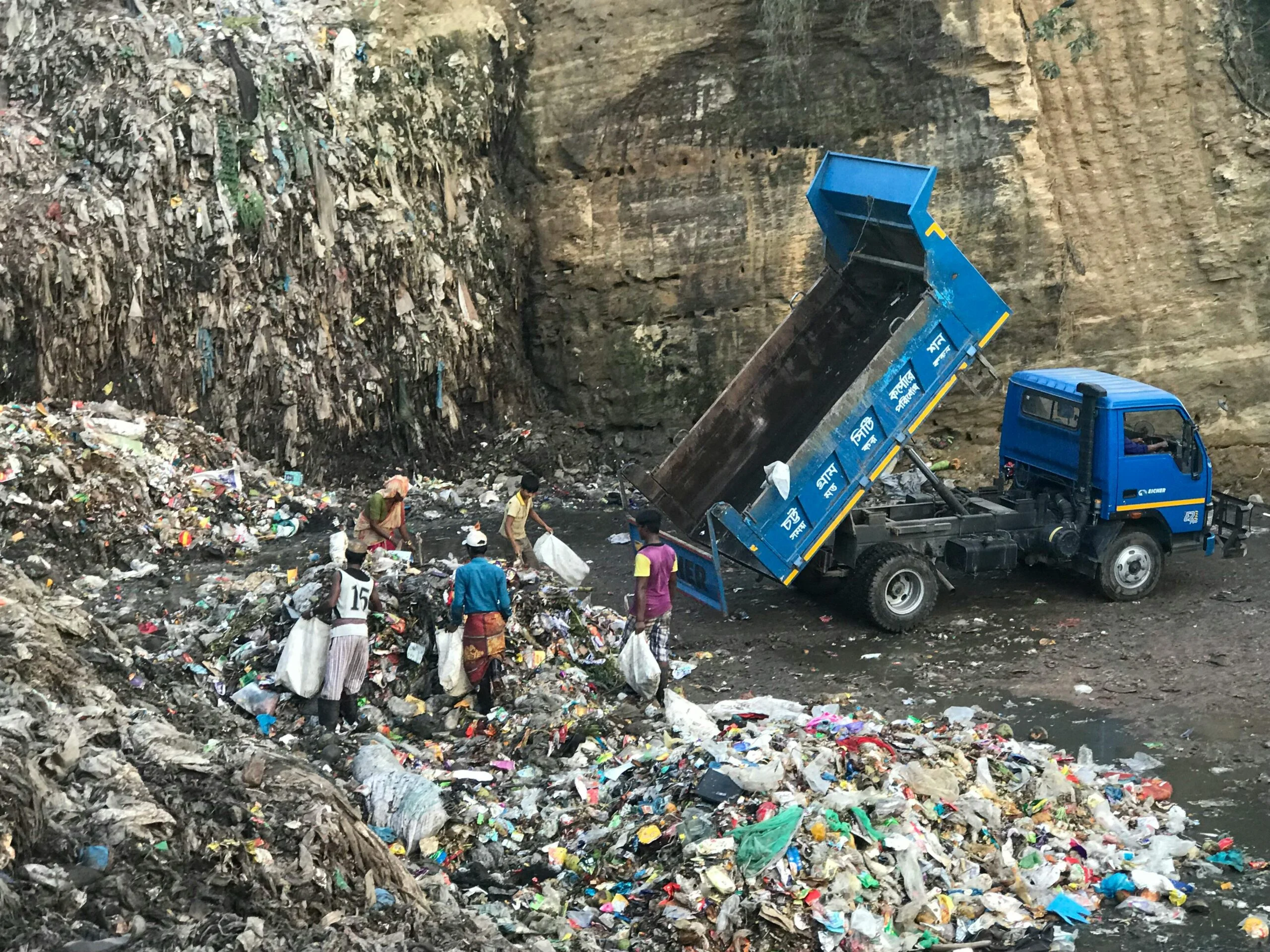 Blue tipper truck unloading rubbish at a landfill beside a cliff, with waste pickers sorting through the heap