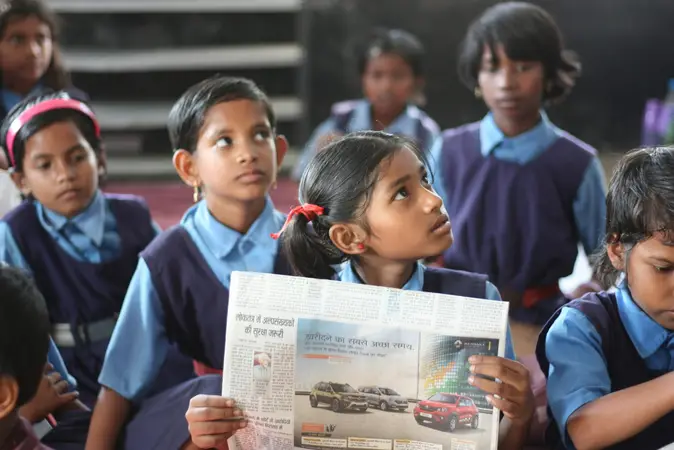 Schoolgirls in blue and purple uniforms sitting on the floor, one holding up a Hindi newspaper