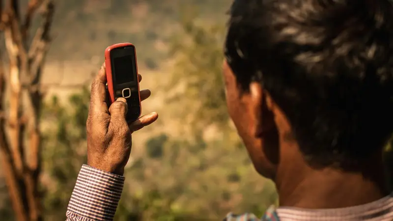Man seen from behind holding a red basic mobile phone against a blurred hillside