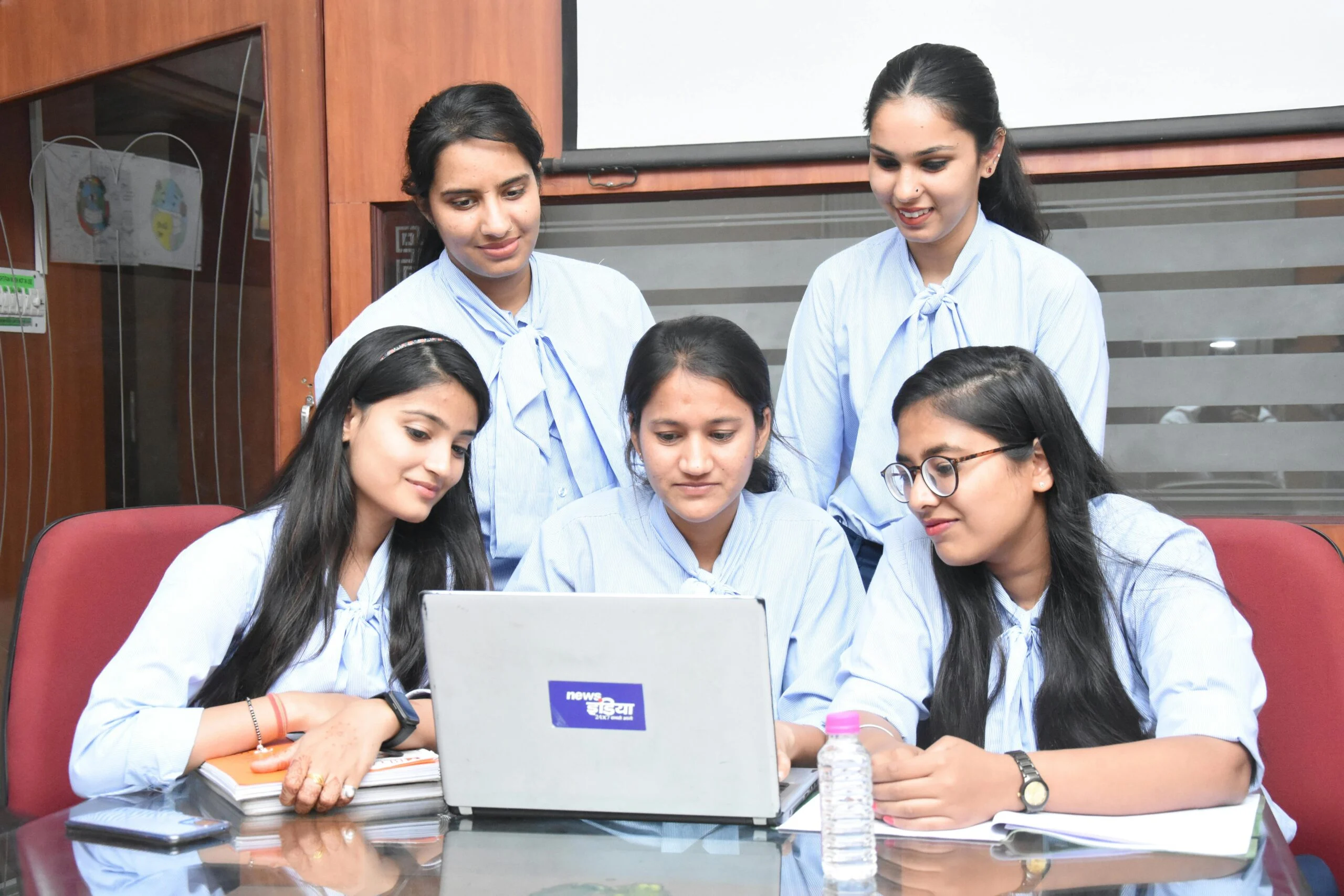 Four young women in light blue uniforms gathered around an open laptop at a table