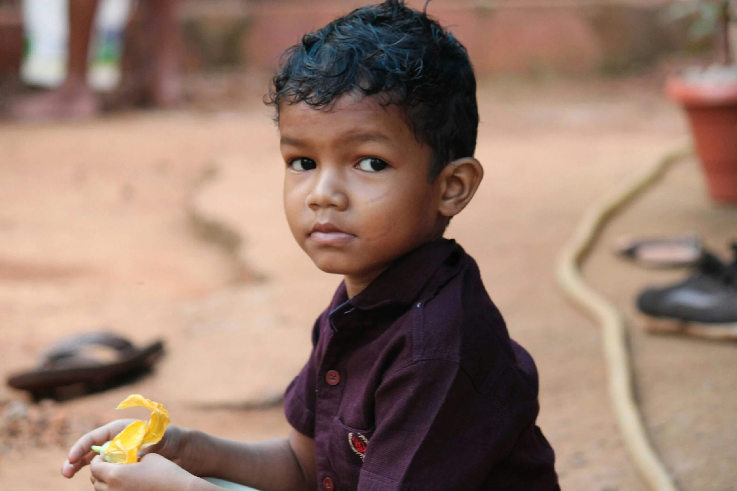 Young boy in a maroon shirt sitting on bare ground holding a yellow wrapper and looking at the camera