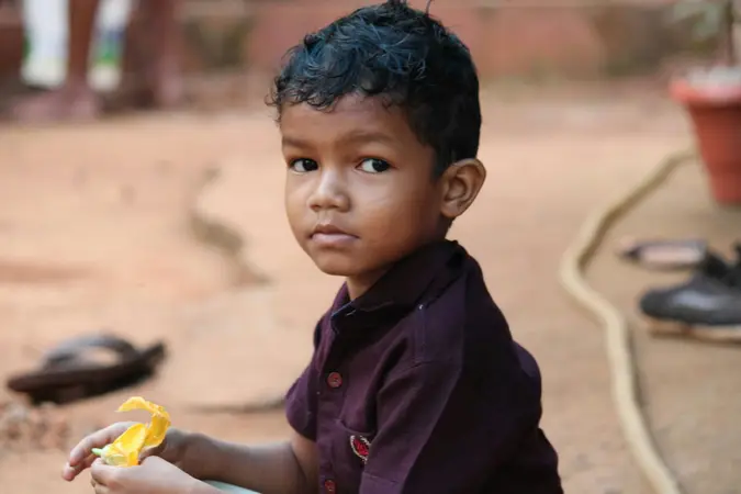 Young boy in a maroon shirt sitting on bare ground holding a yellow wrapper and looking at the camera