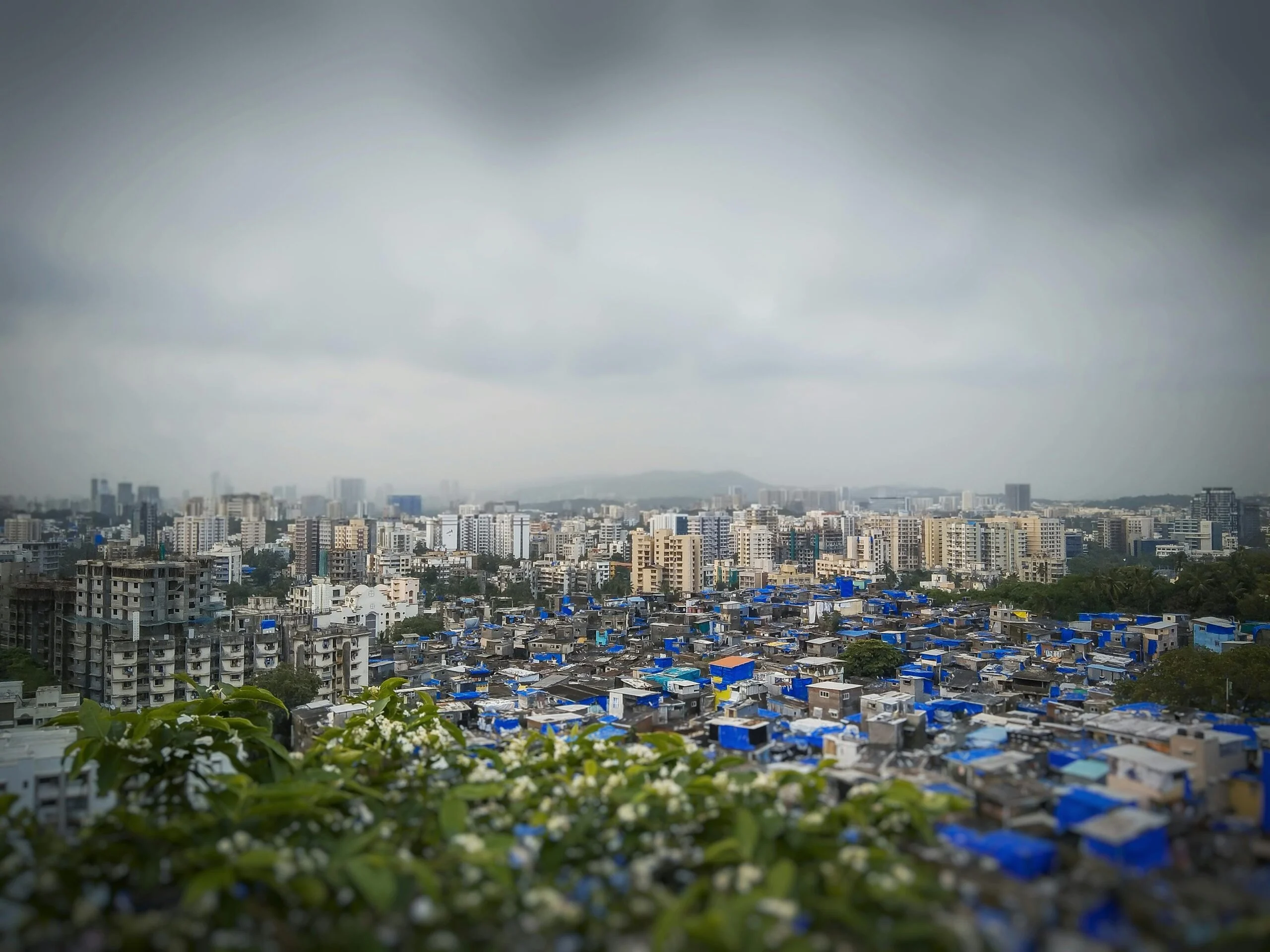 View over Mumbai with blue tarpaulin-covered Dharavi rooftops in the foreground and high-rise towers beyond under a grey sky