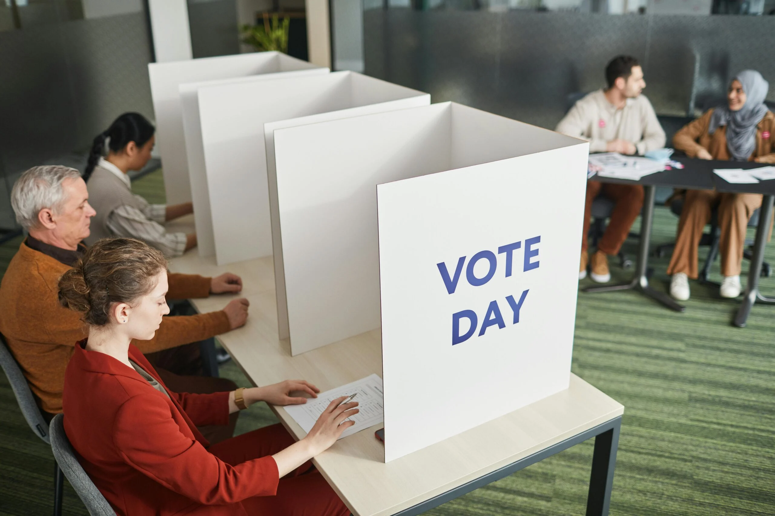 Voters filling forms at privacy booths, one marked VOTE DAY, with people seated at a table in the background