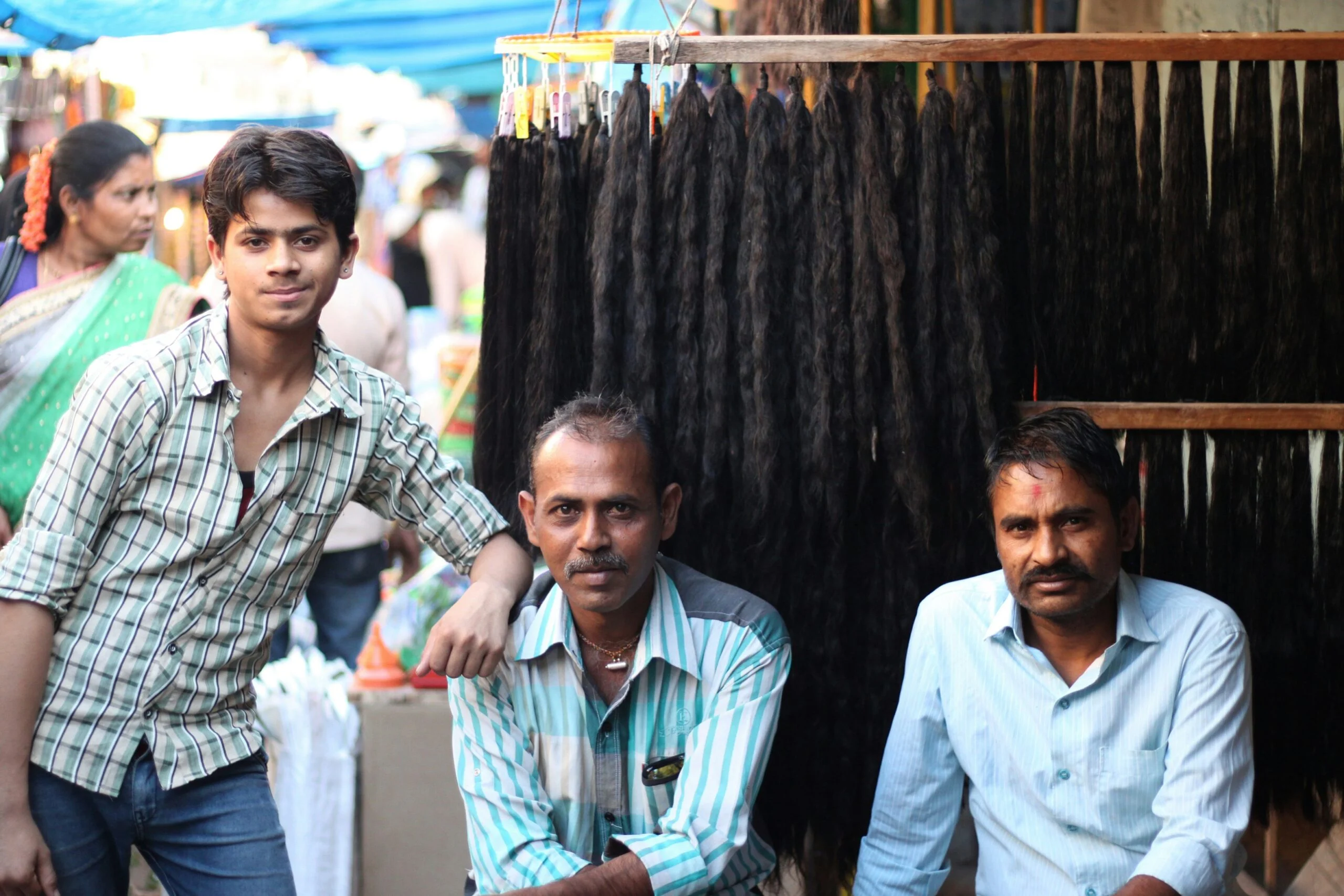 Two seated men and a standing teenage boy in front of hanging bundles of long black hair at a market stall