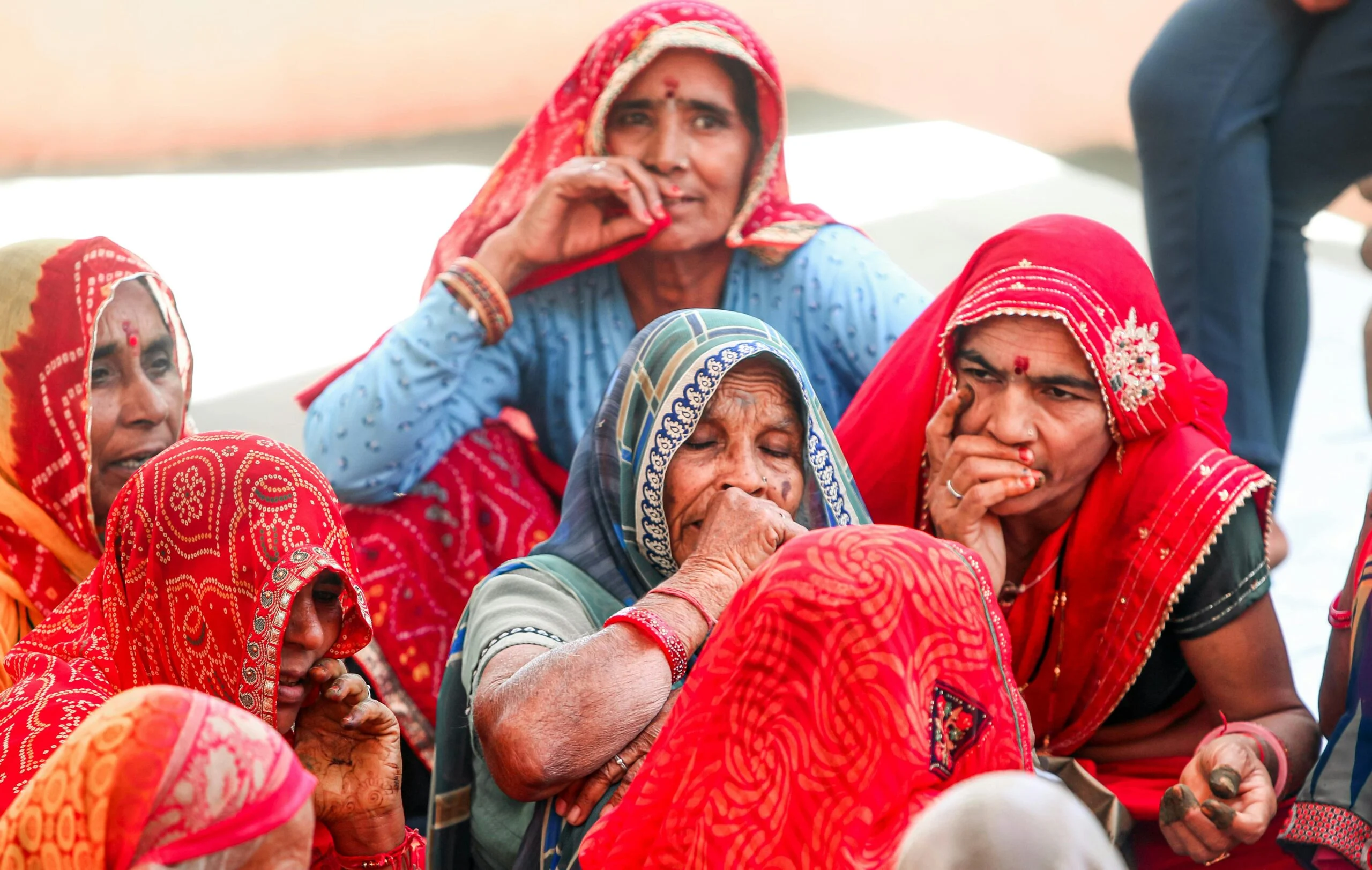 Group of women in red and blue saris sitting close together, several with a hand raised to their face