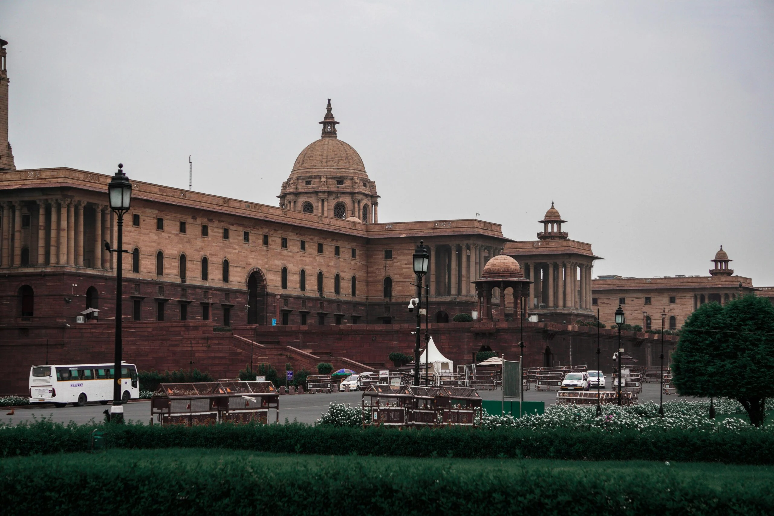 Red sandstone government buildings with a dome behind street lamps, a white bus and barricades