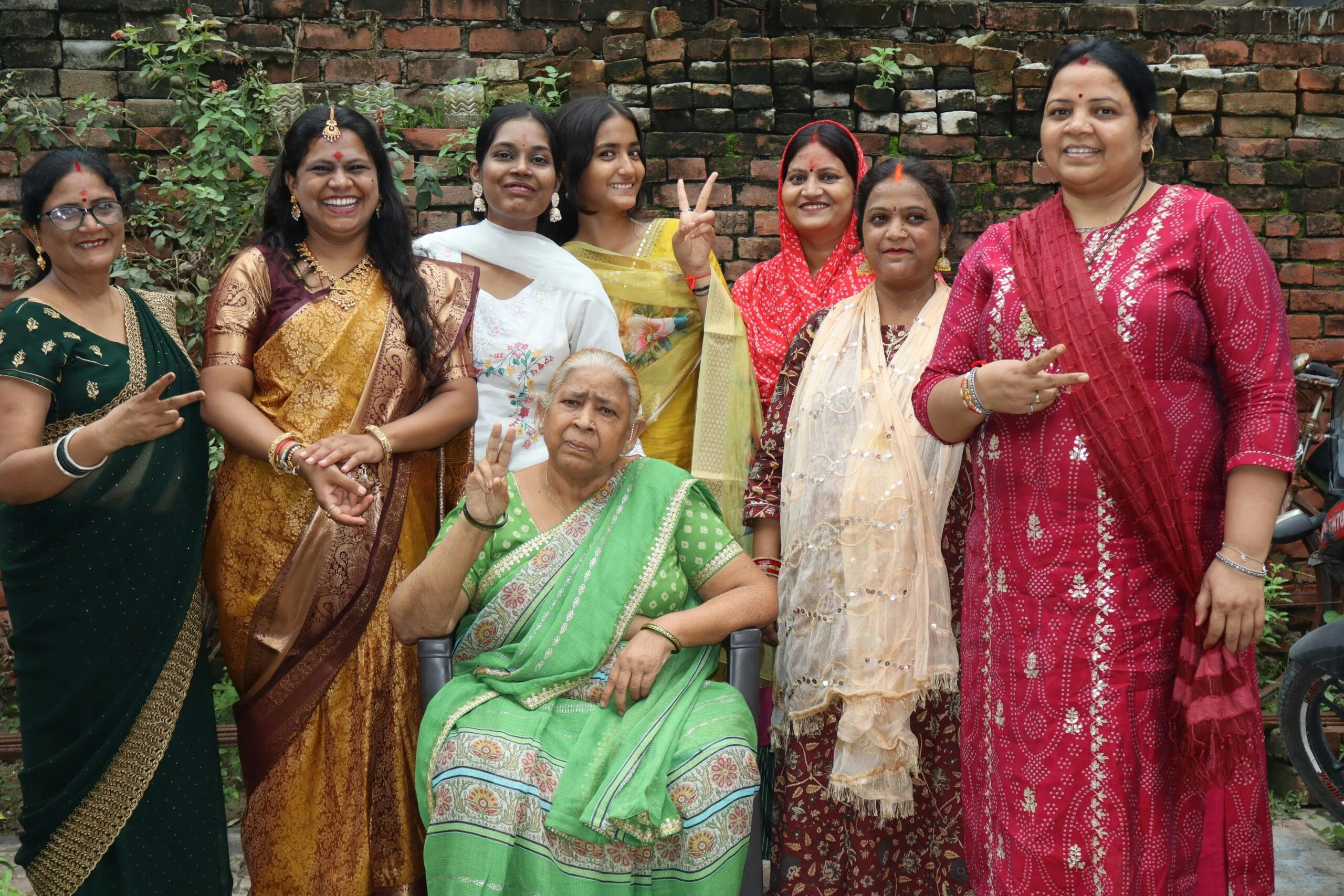 Group of smiling women in colourful saris posing with peace signs in front of a brick wall