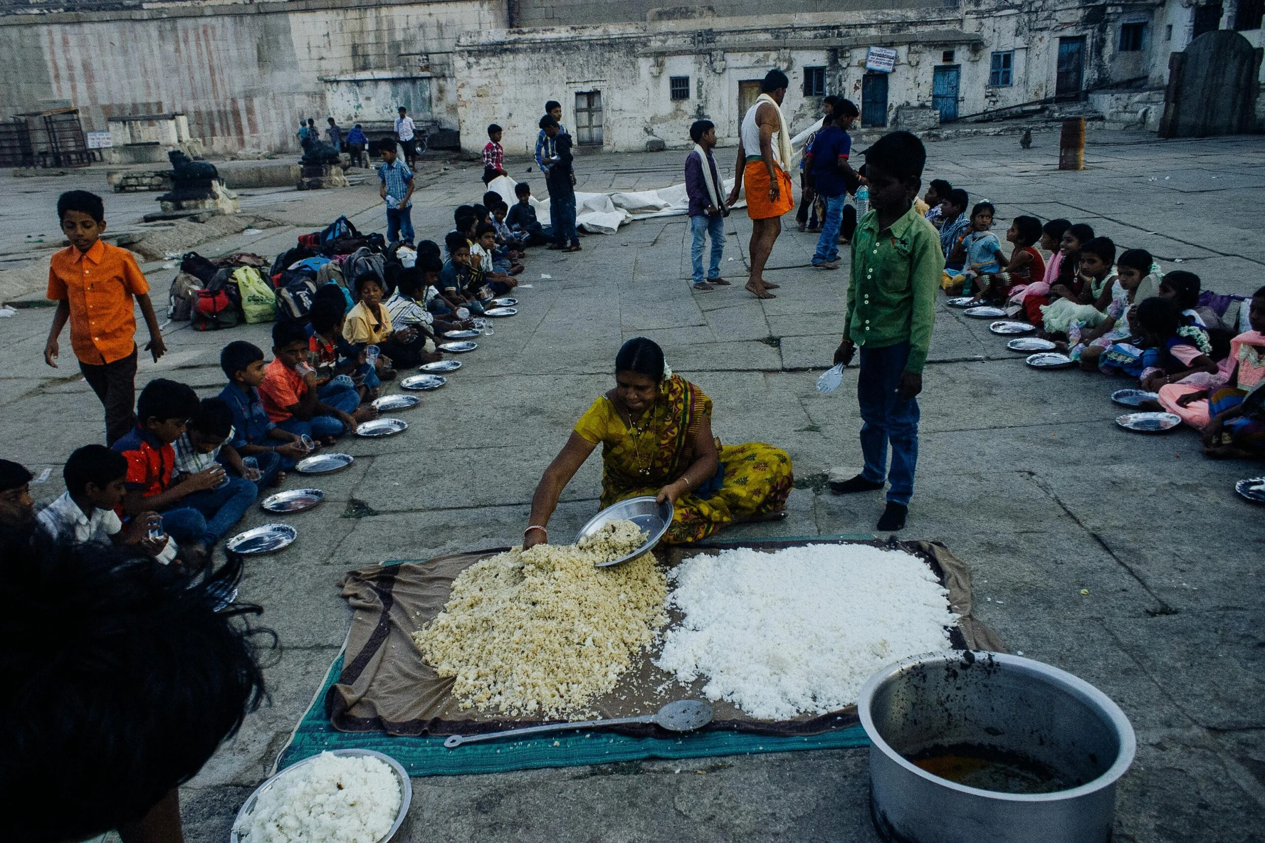 Woman serving rice from a mat to children seated in rows with plates on a stone courtyard