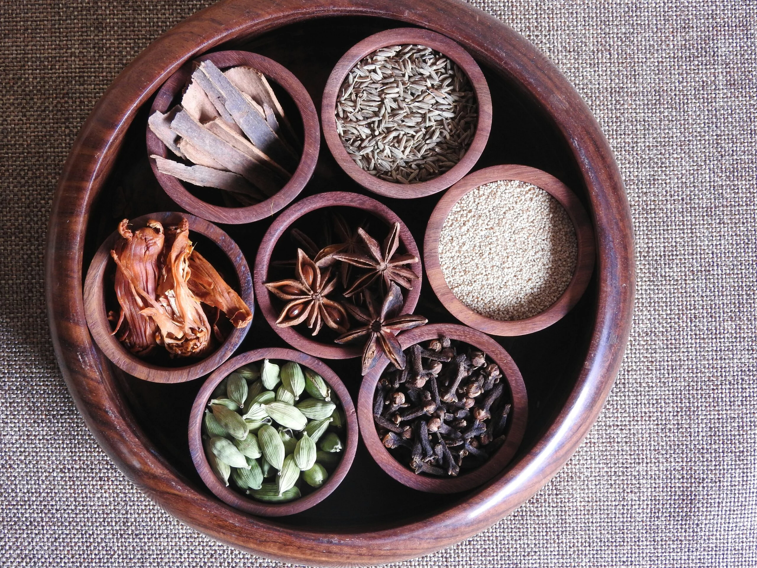 Wooden bowl holding smaller bowls of cinnamon, cumin, star anise, mace, cardamom and cloves
