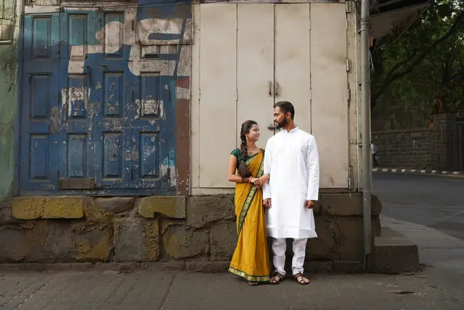 Young couple in a yellow sari and white kurta standing side by side in front of weathered blue and cream wooden shutters