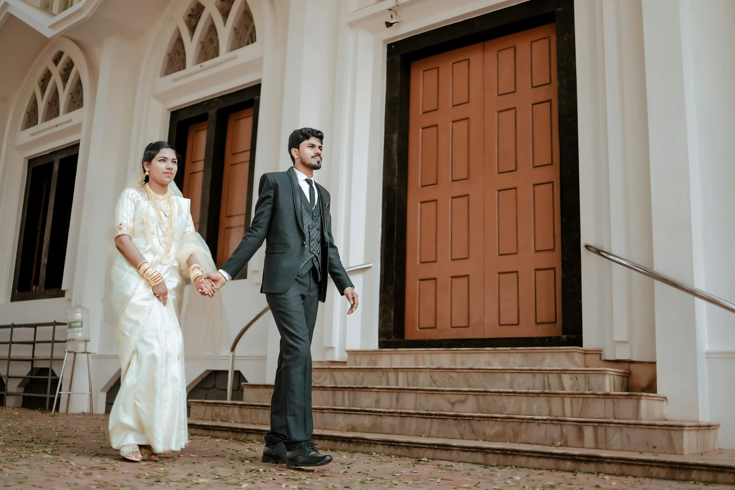 Bride in a white sari and groom in a dark suit holding hands and walking past wooden doors of a white building