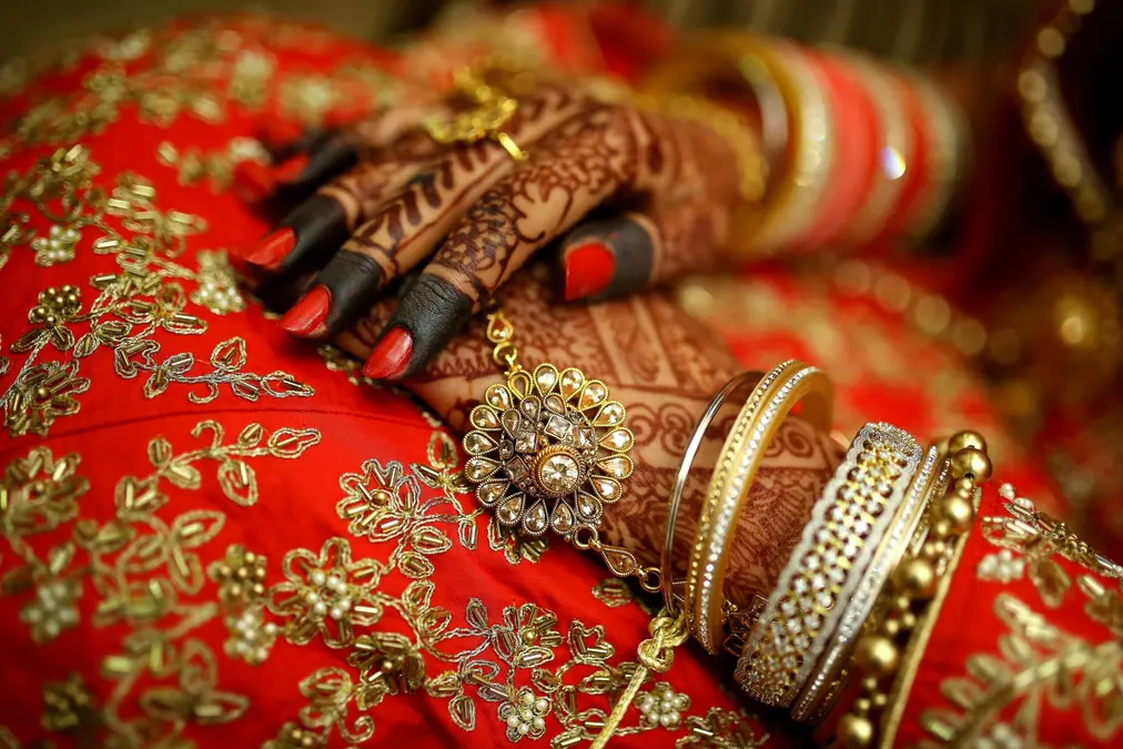 Bride's hands with henna, red nail polish and gold bangles resting on red embroidered fabric
