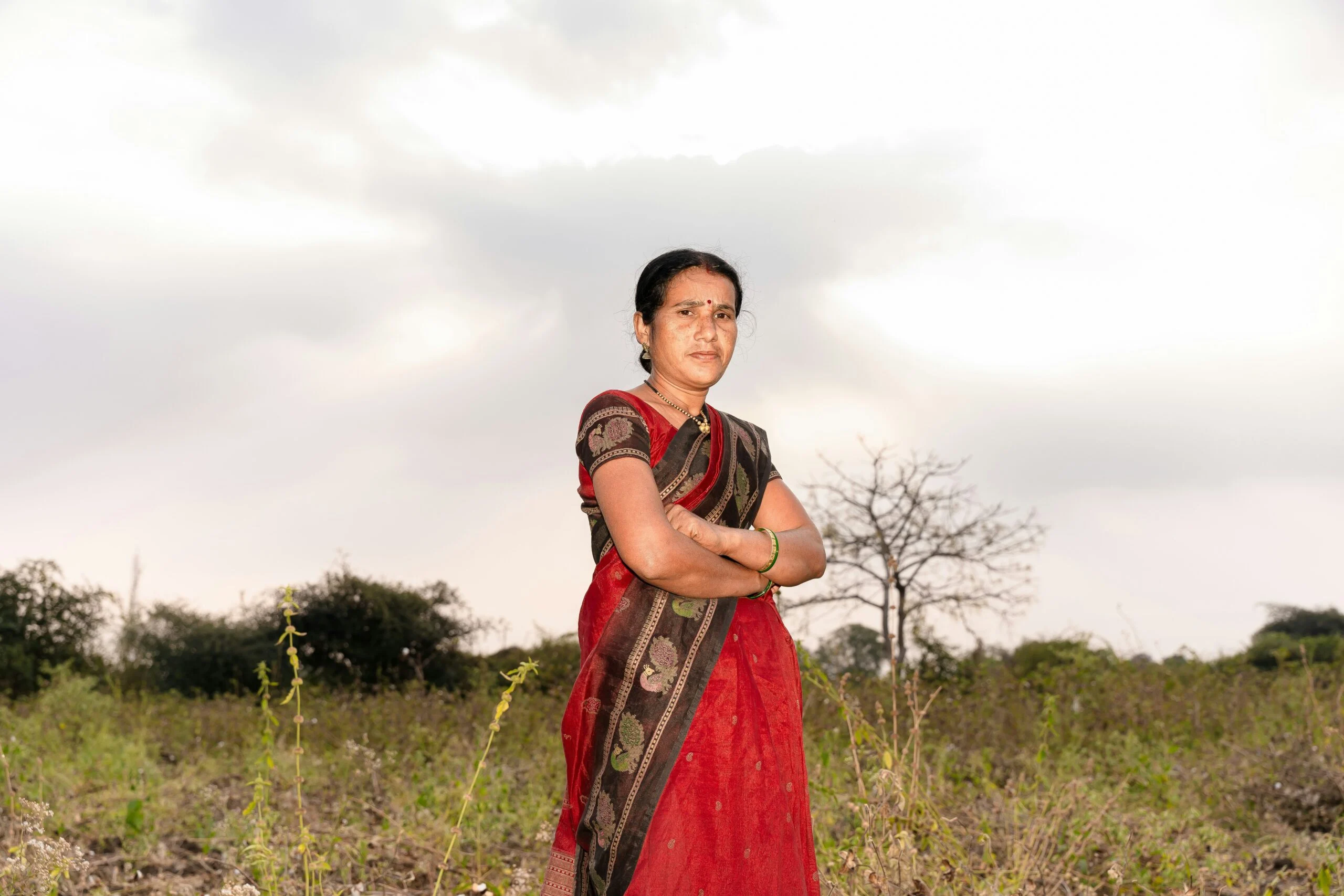 Woman in a red sari with arms crossed standing in a scrubby field under an overcast sky