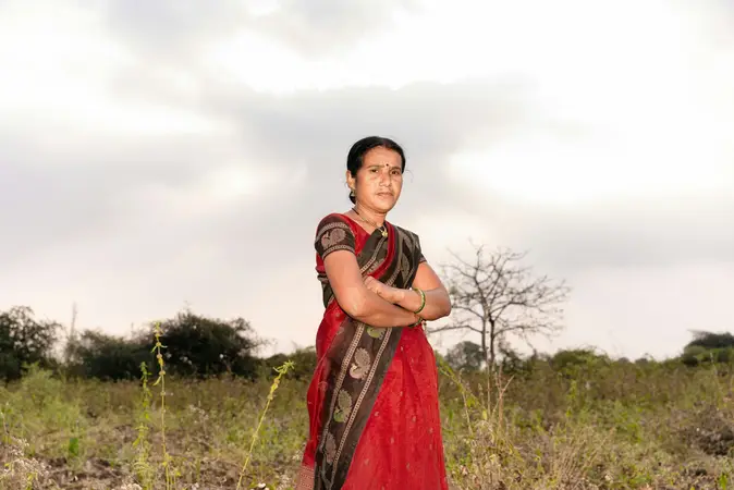 Woman in a red sari with arms crossed standing in a scrubby field under an overcast sky