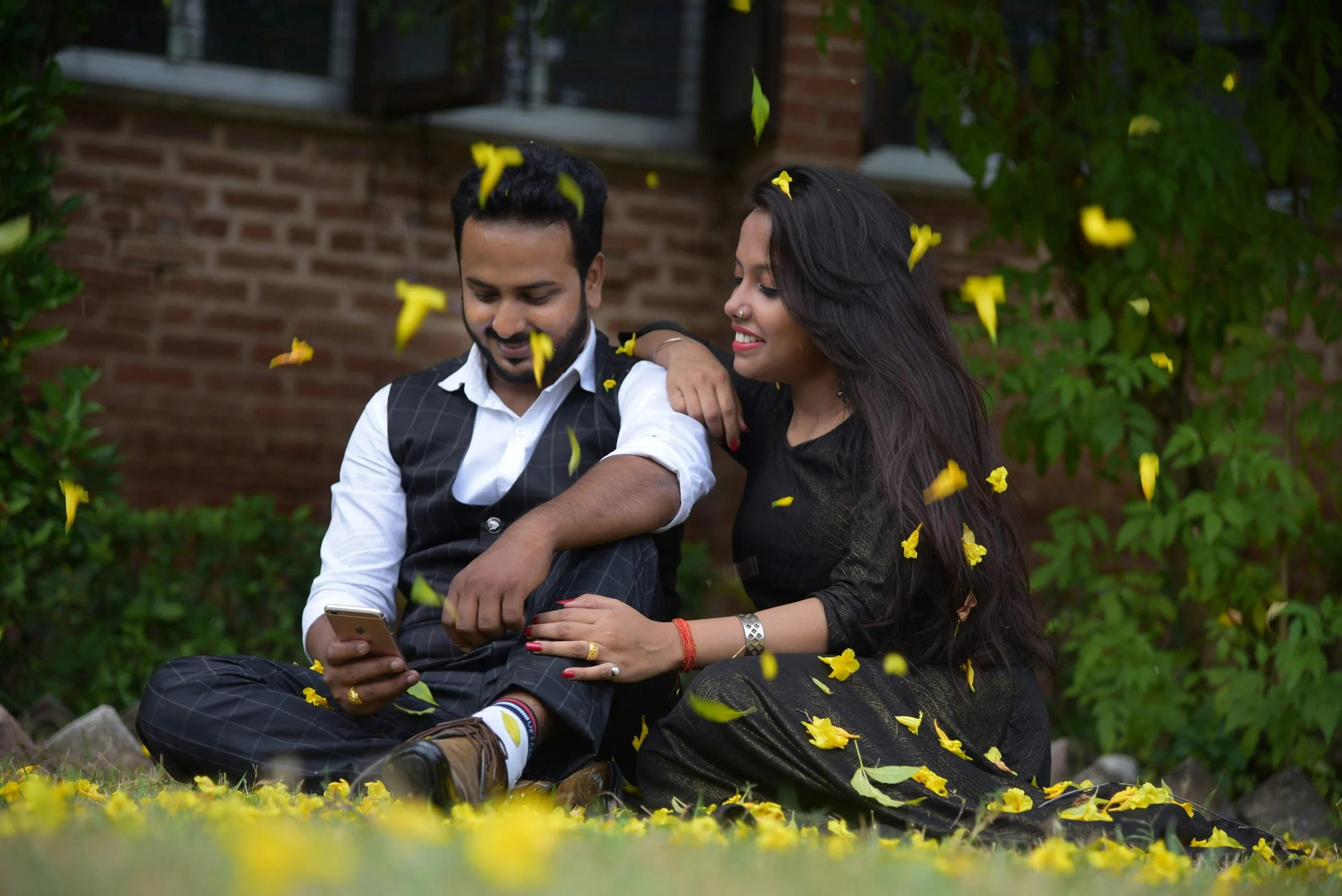 Young couple sitting on grass looking at a phone while yellow flower petals fall around them
