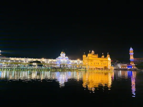 Golden Temple in Amritsar lit up at night, with its golden reflection on the dark water and a blue-lit minaret beside it