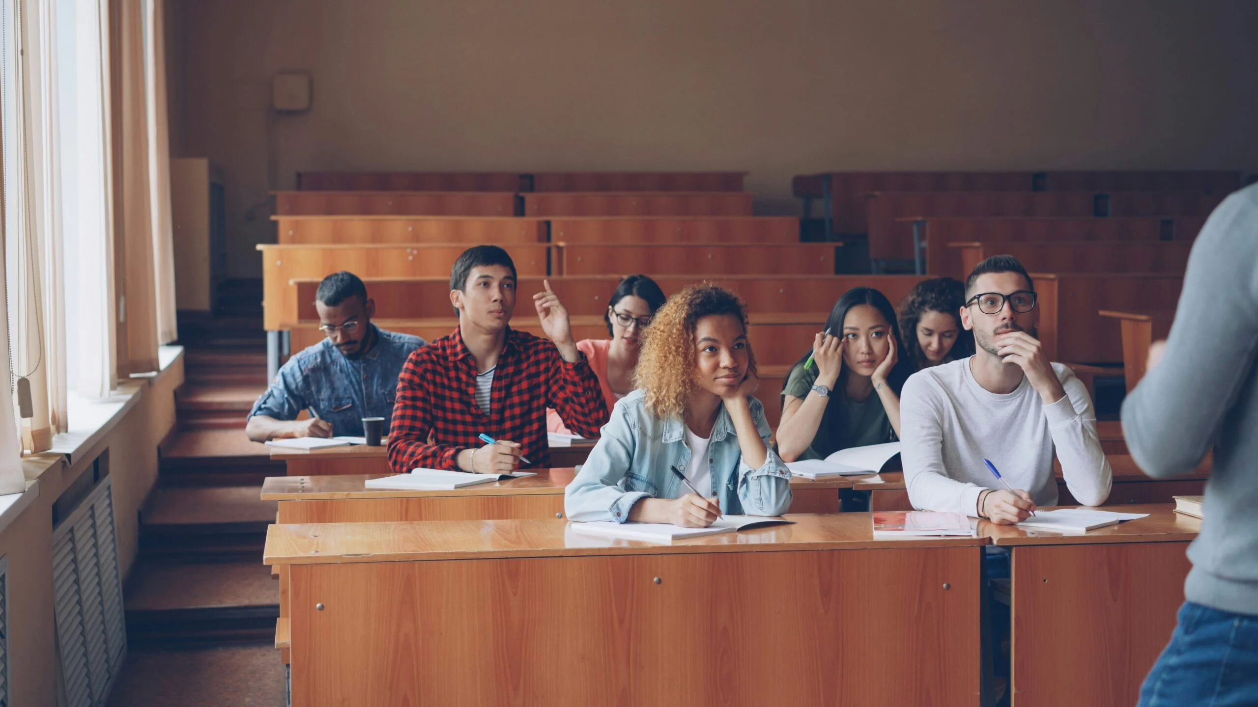 Students seated at wooden desks in a lecture hall, some raising hands, listening to a teacher