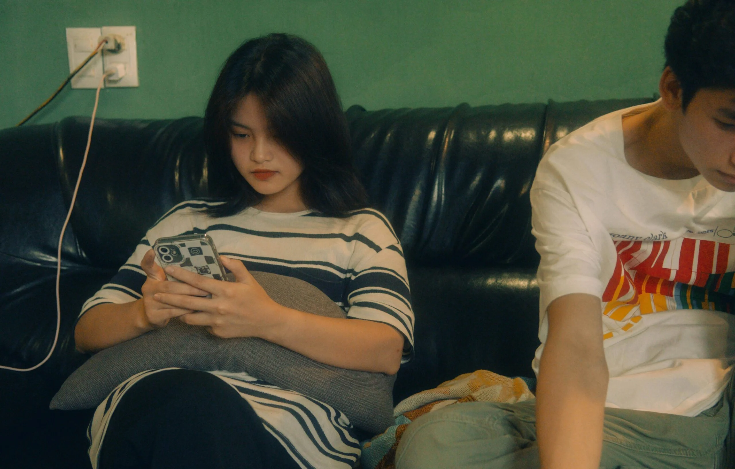 Teenage girl in a striped top looking at her phone on a black leather sofa beside a boy
