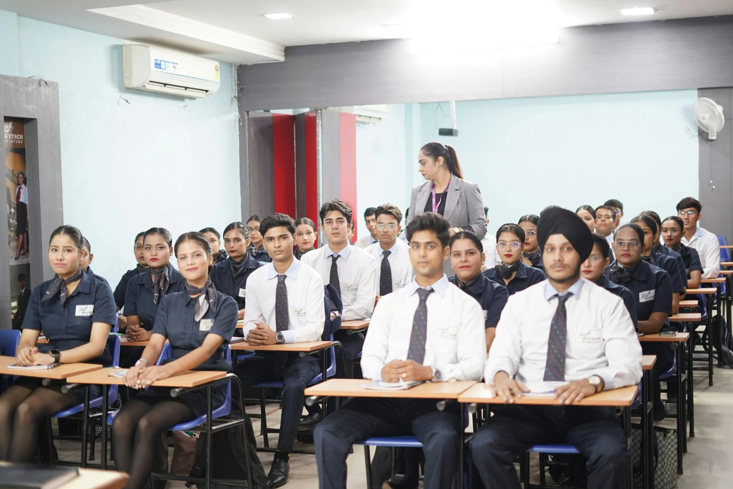 Trainees in dark uniforms and ties sitting at desks in a classroom while an instructor stands at the back
