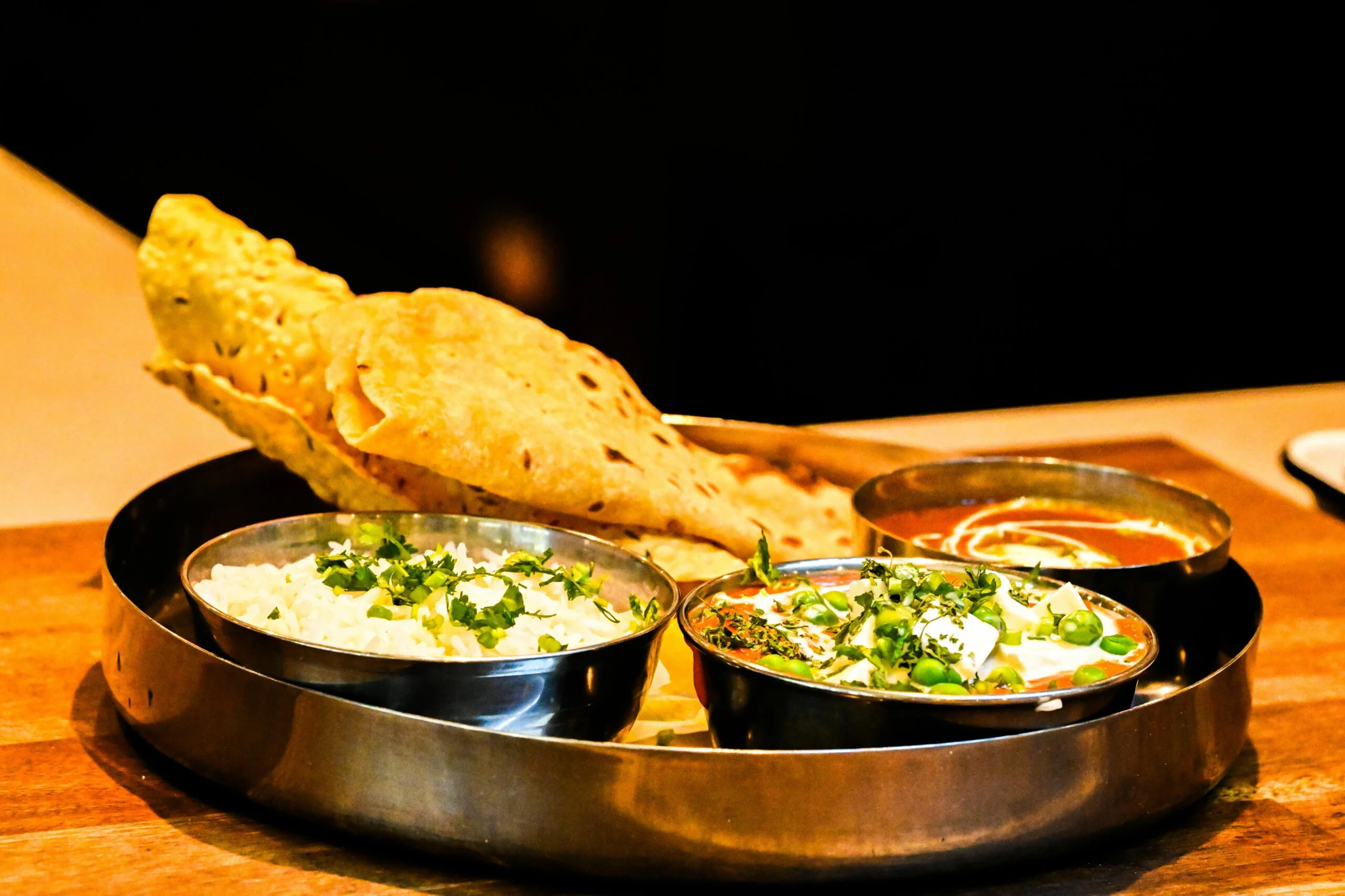Steel thali with papad, rice and two bowls of vegetable curries garnished with herbs on a wooden table