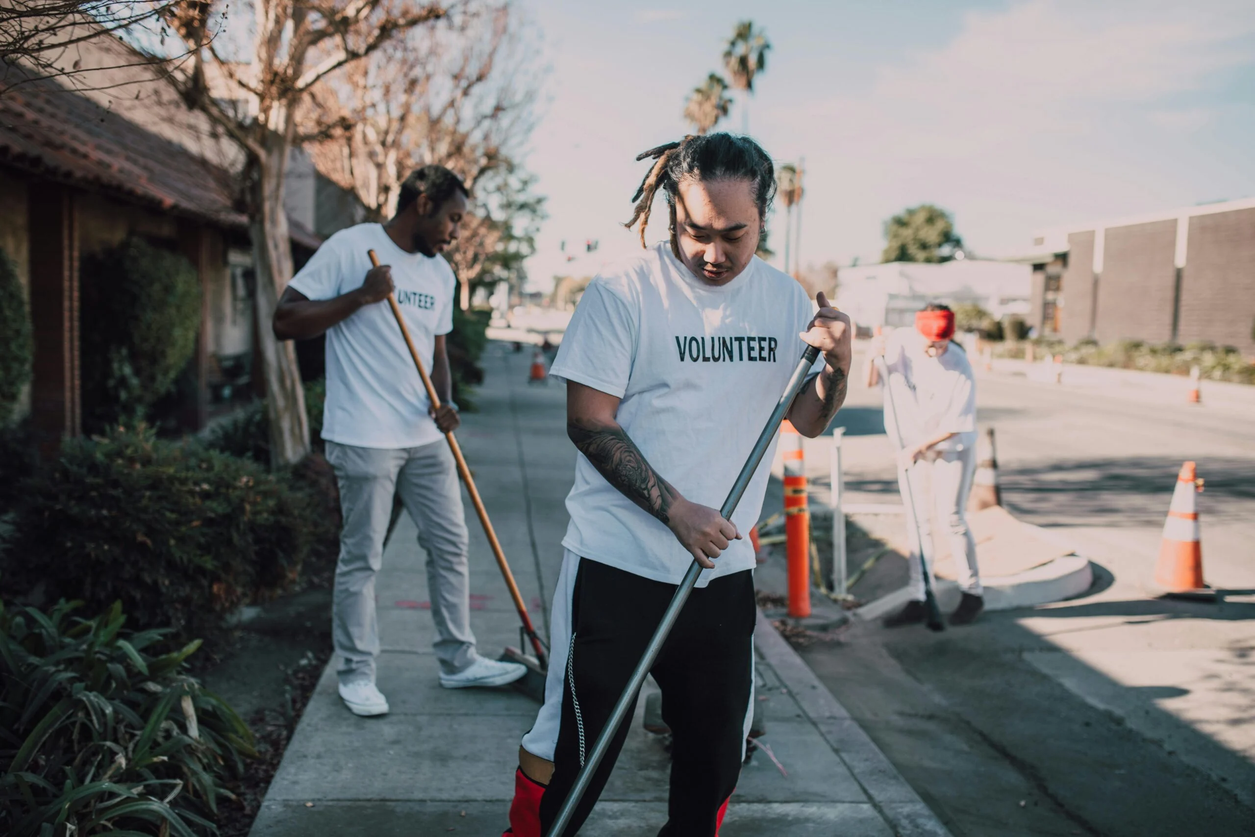Two volunteers in white Volunteer T-shirts sweeping a sidewalk with brooms near orange traffic cones