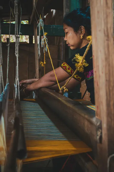 Woman weaving on a wooden handloom in a dim workshop, hands on the loom beam and threads hanging beside her