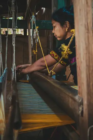 Woman weaving on a wooden handloom in a dim workshop, hands on the loom beam and threads hanging beside her