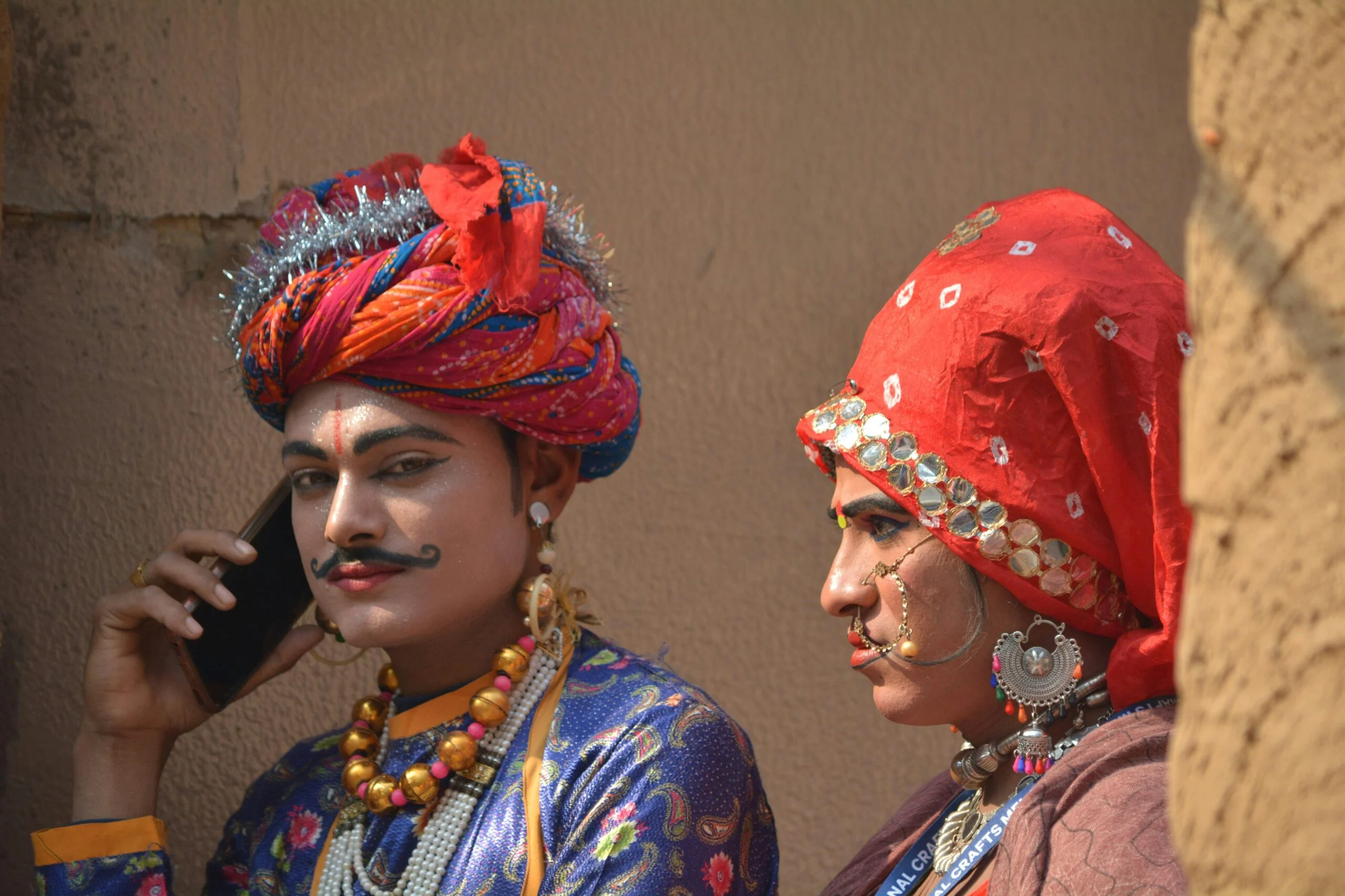 Two people in bright turbans and embroidered costumes, one speaking on a mobile phone, against a mud wall