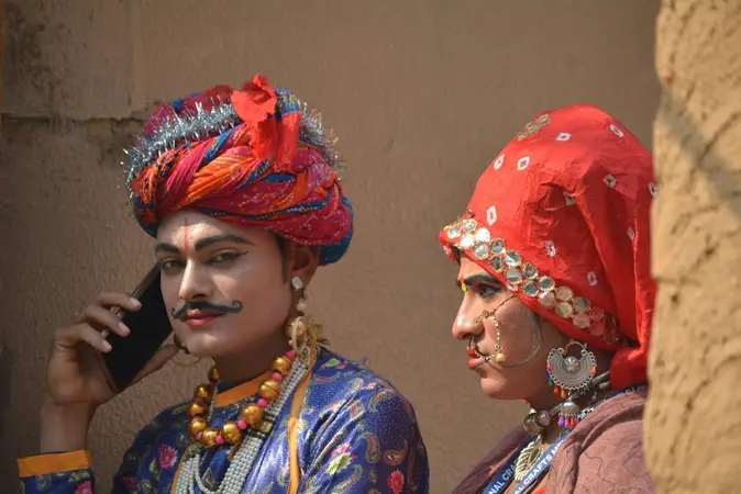 Two people in bright turbans and embroidered costumes, one speaking on a mobile phone, against a mud wall