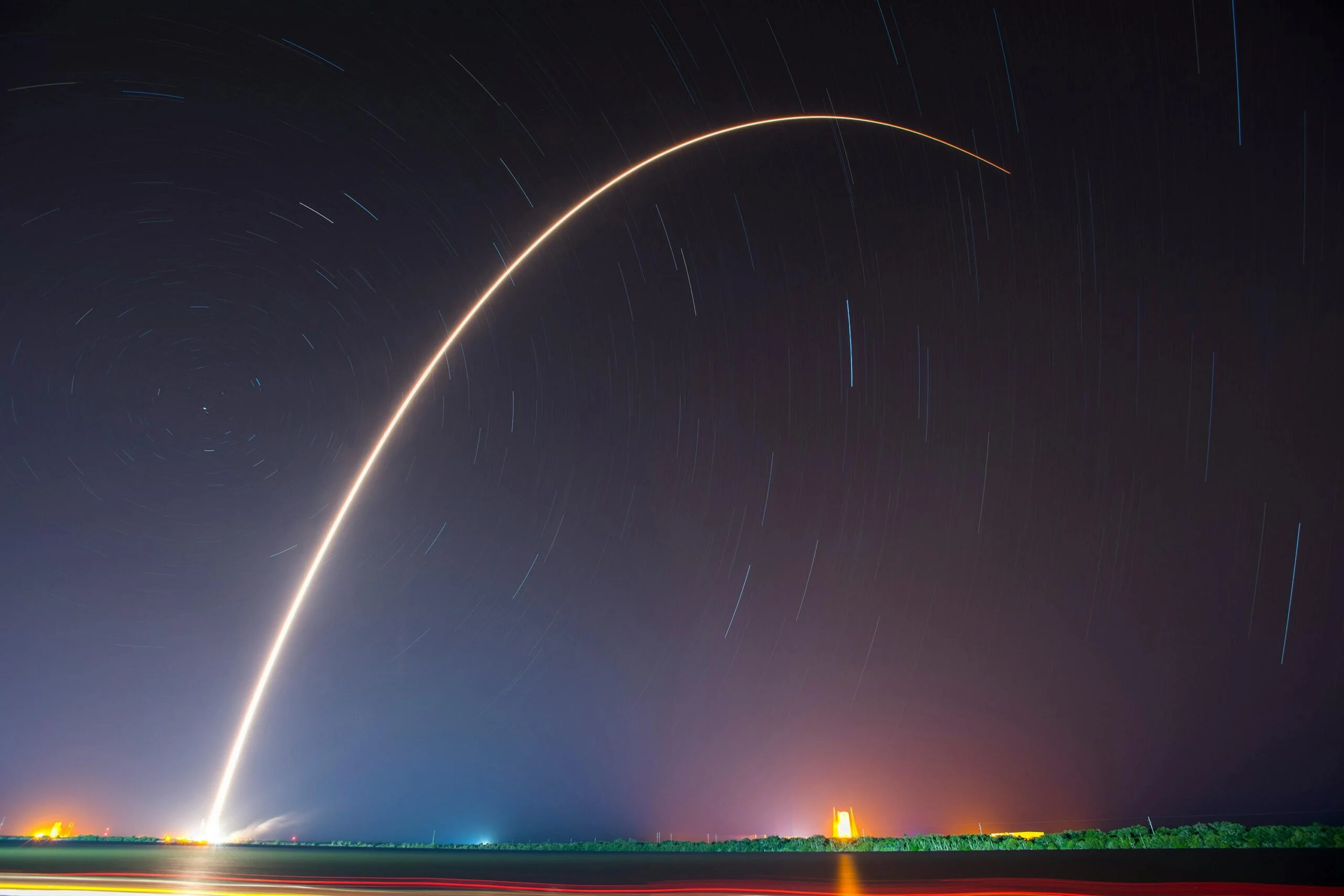 Long-exposure night photo of a rocket launch leaving a bright curved arc of light across a starry sky