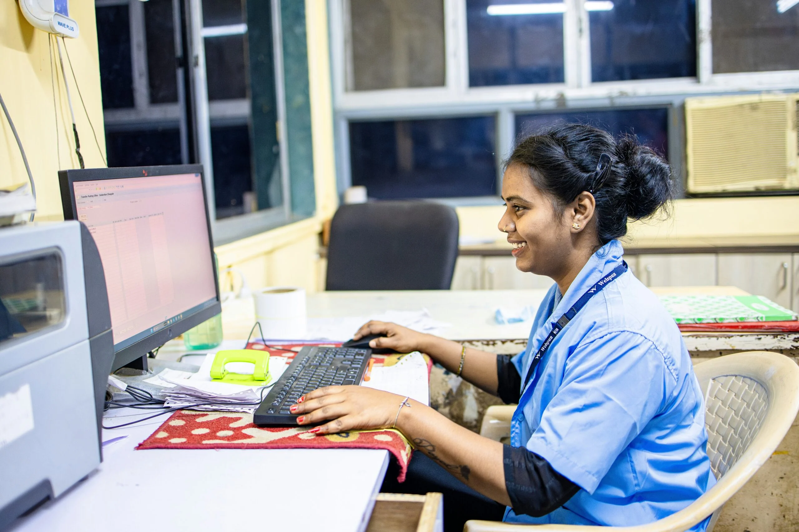 Smiling woman in a light blue uniform with an ID lanyard working at a computer keyboard in an office