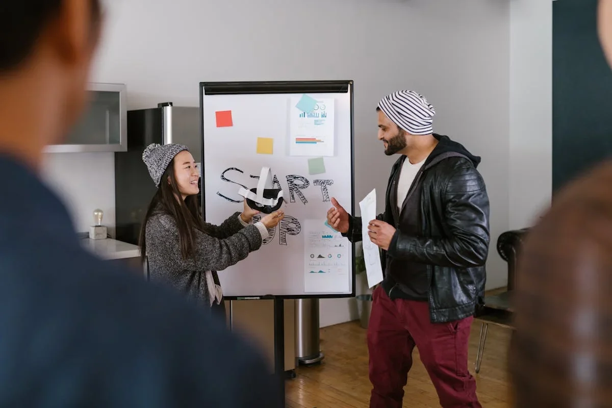 Man in a beanie and leather jacket and a woman in a grey beanie talking beside a whiteboard with the word START on it