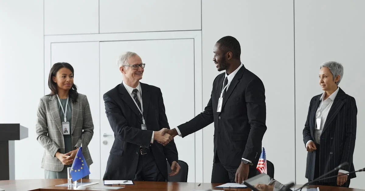 Two men in suits shaking hands at a conference table with an EU flag and a small US flag, two colleagues looking on