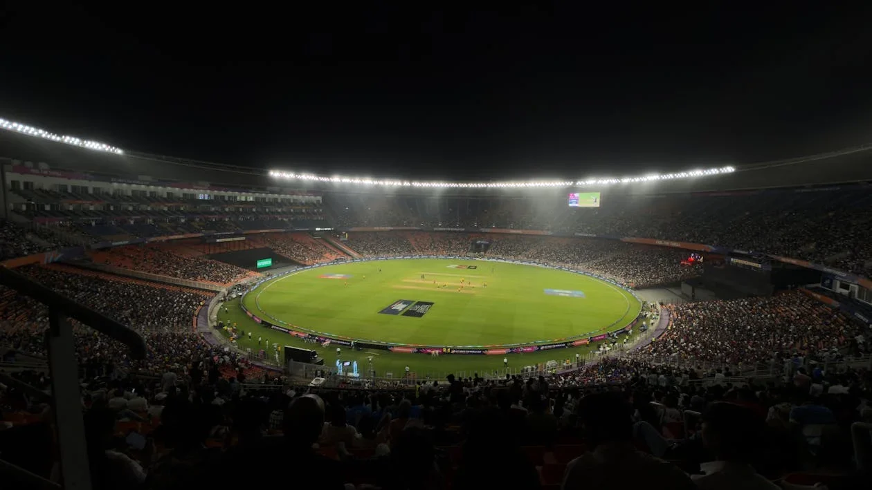 Floodlit cricket stadium at night with a packed crowd around a green field