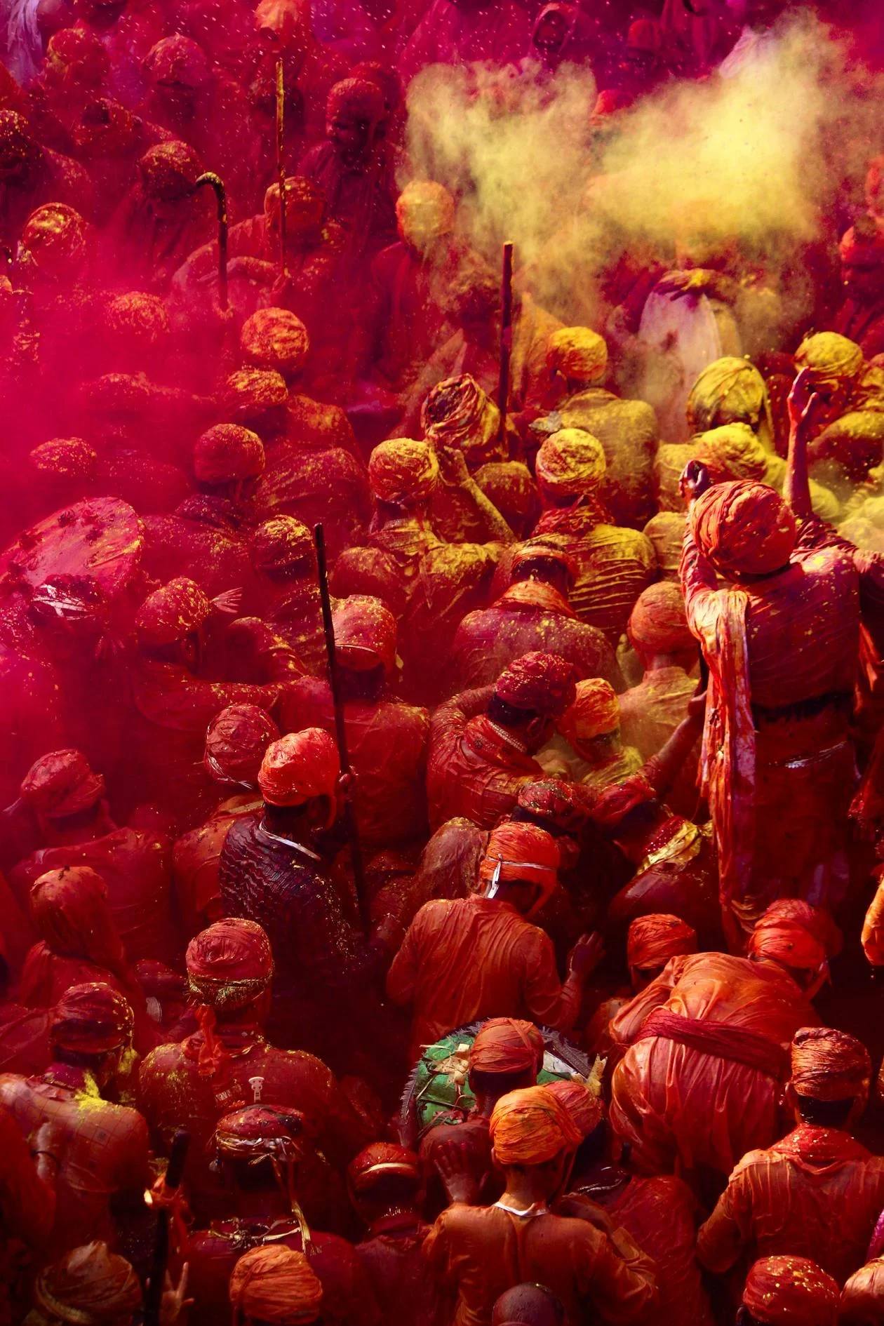 Crowd covered in red and orange powder wearing turbans, with yellow powder clouds, seen from above