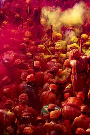 Crowd covered in red and orange powder wearing turbans, with yellow powder clouds, seen from above