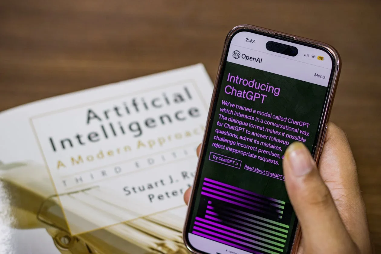 Hand holding a phone showing the ChatGPT introduction page, resting on an Artificial Intelligence textbook