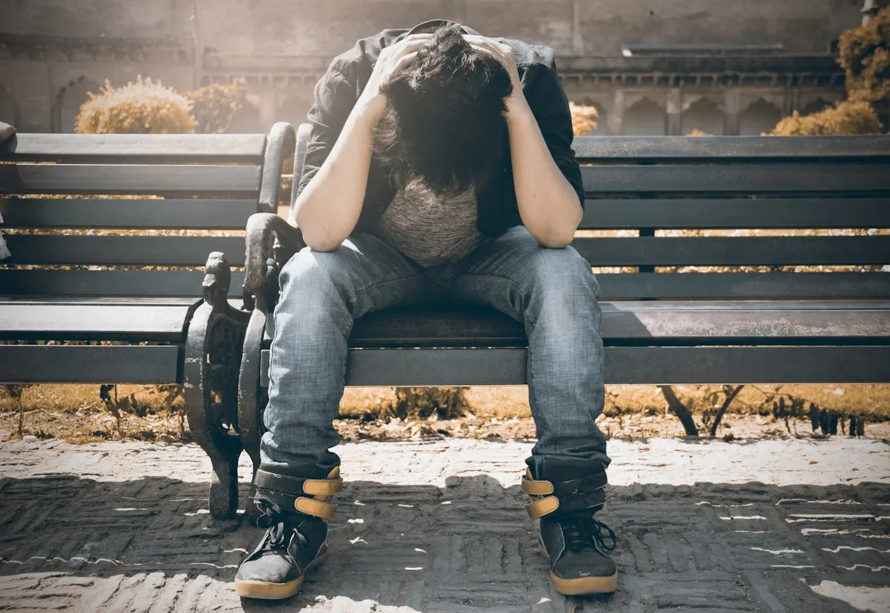Person sitting on a park bench with head bowed in their hands, face hidden