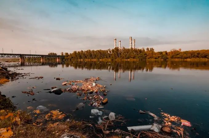 Polluted river with floating plastic and debris in the foreground and chimney stacks beyond the trees