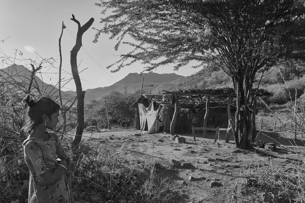 Black and white image of a girl standing in a dry village clearing near a thatched shelter, with hills behind