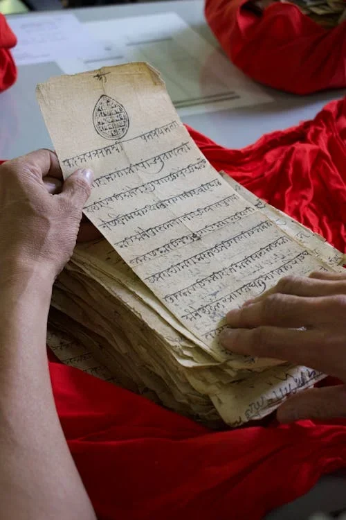 Hands holding a stack of old handwritten manuscript pages in Indic script over a red cloth