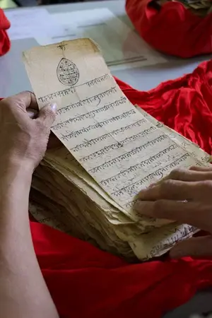 Hands holding a stack of old handwritten manuscript pages in Indic script over a red cloth