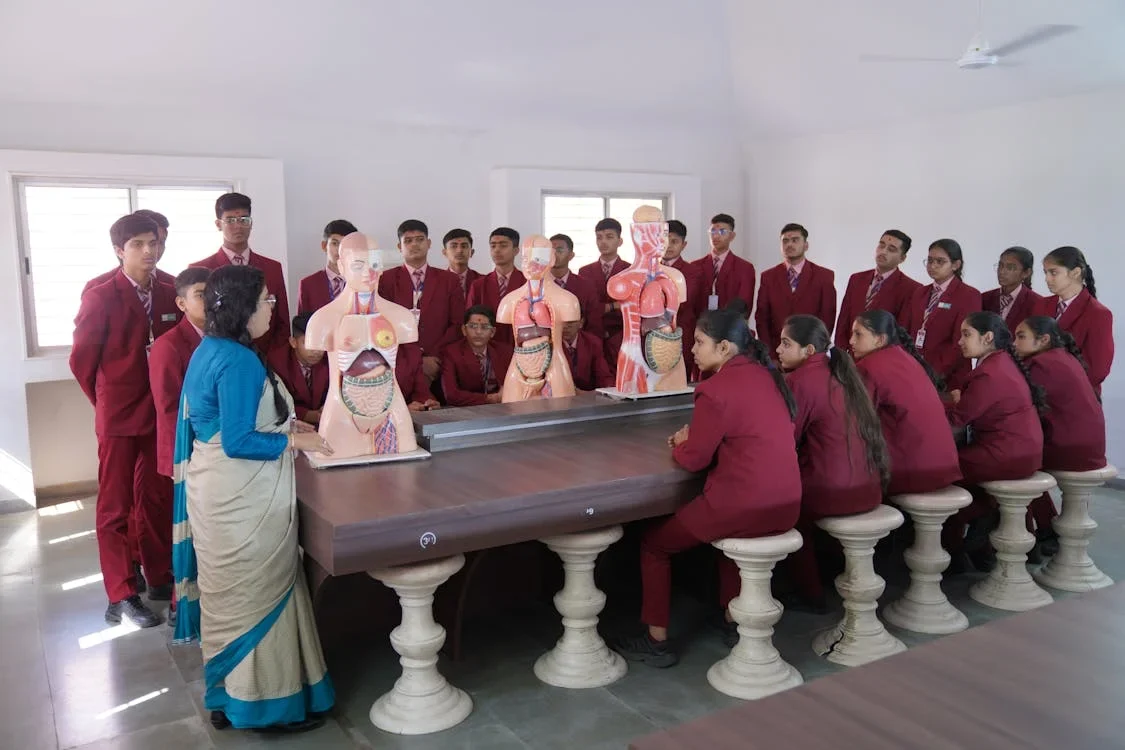 Teacher in a sari showing anatomy models to students in maroon uniforms seated and standing around a long table