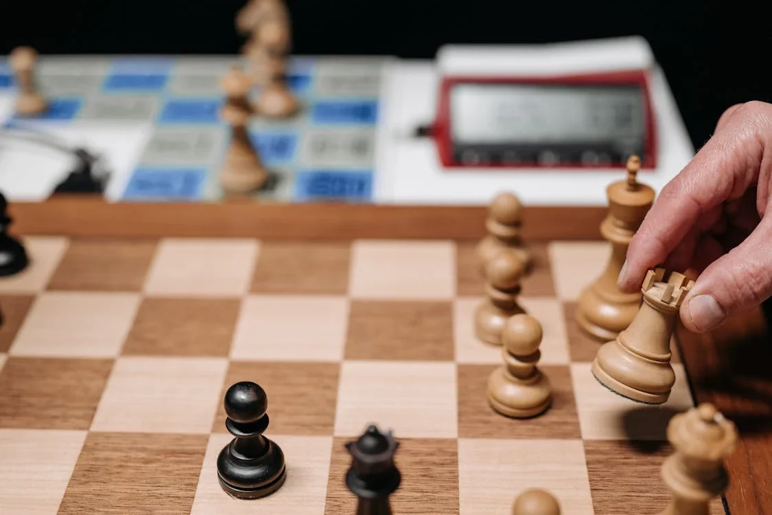 Hand moving a white rook on a wooden chess board with a digital chess clock in the background