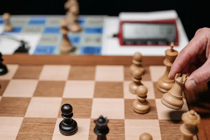 Hand moving a white rook on a wooden chess board with a digital chess clock in the background